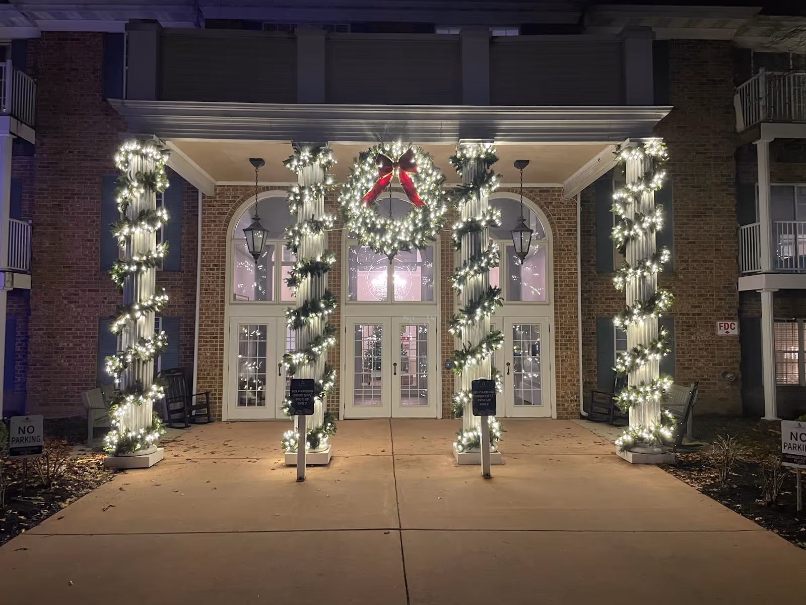 Night view of a brick building entrance decorated with illuminated garlands wrapped around columns and a large wreath above the doors.