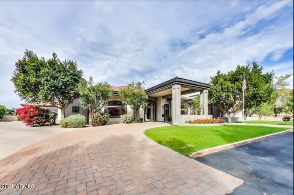 Front entrance of a single-story assisted living building with a covered porte-cochere, landscaped lawn, trees, and an American flag.