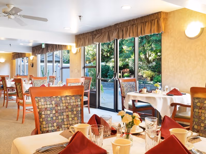 Dining room with set tables, upholstered chairs, and glass doors opening to a garden.