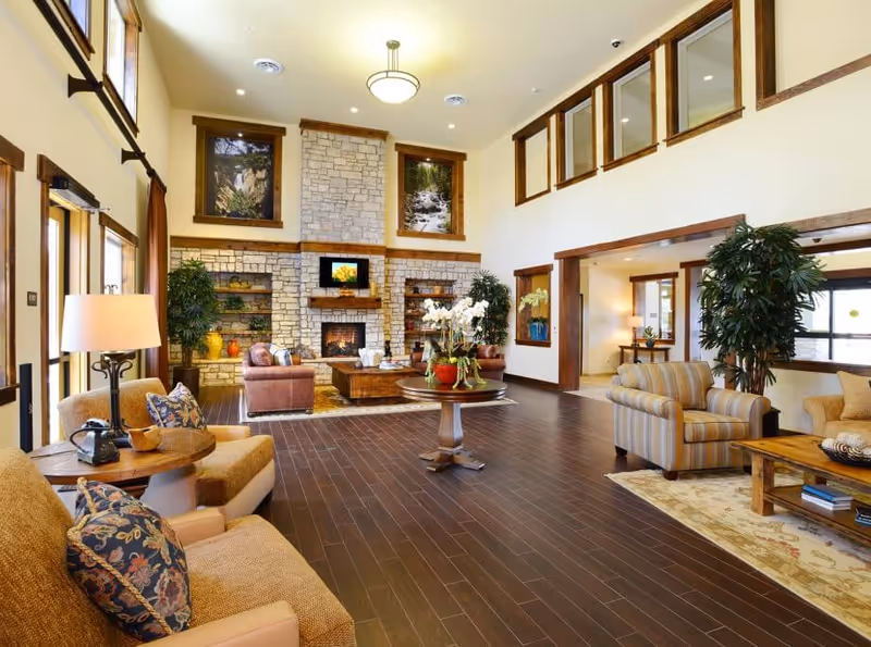 A bright, spacious senior living facility living room with a stone fireplace, multiple seating areas, and dark wood flooring.