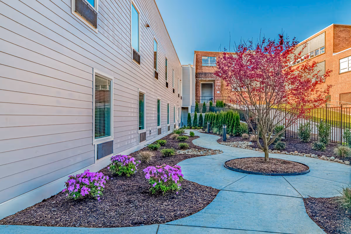 Curved paved courtyard with landscaped flower beds and a small tree alongside a senior living building exterior.