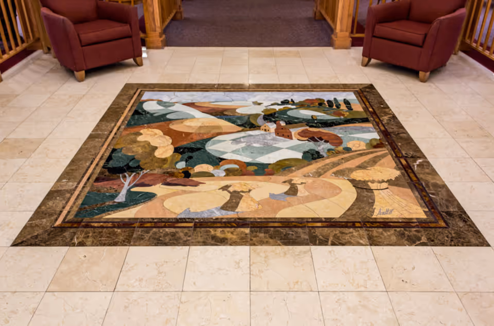 A decorative tile floor with an artistic mosaic depicting a countryside landscape with trees, fields, and buildings, bordered by darker tiles. Two red armchairs are positioned on either side of the mosaic, with wooden railings and carpeted flooring visible in the background.