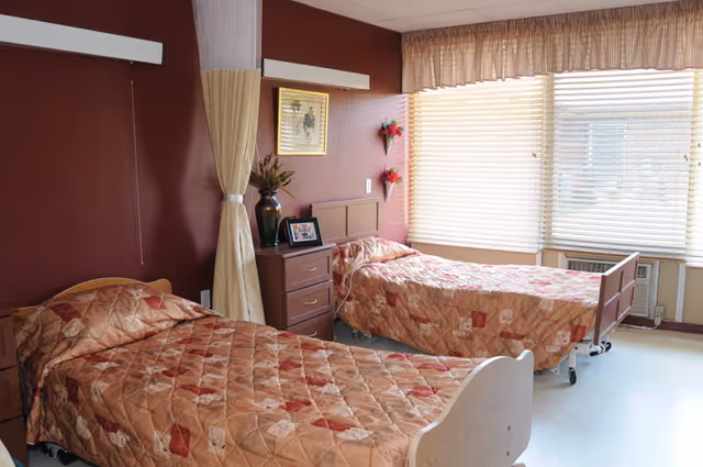 Two twin nursing beds with patterned bedspreads in a shared, sunlit patient room with maroon walls, a bedside dresser, and window blinds.