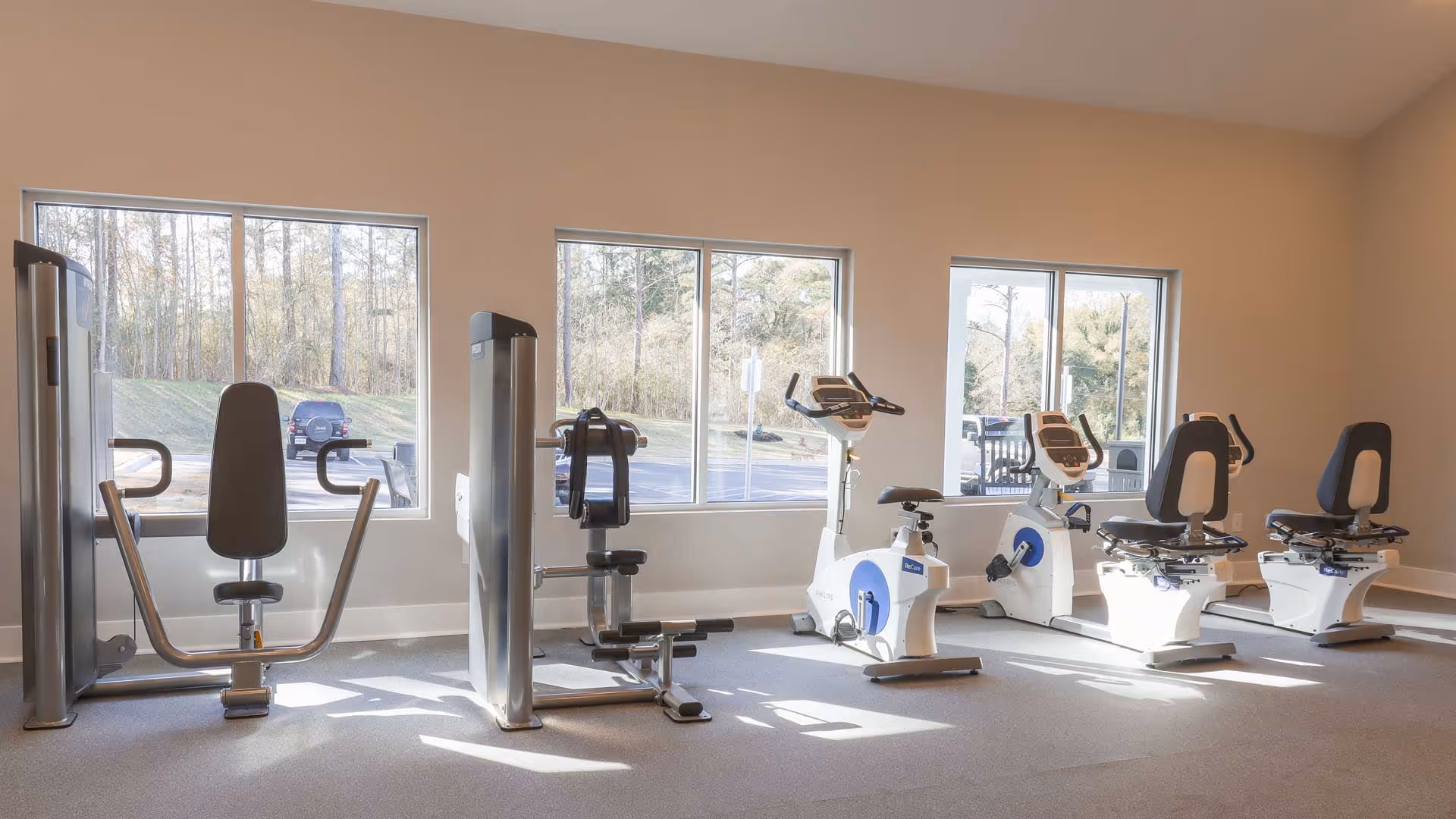 A bright exercise room with large windows showing an outdoor view. The room contains several pieces of fitness equipment including two stationary recumbent bikes and two strength training machines. The floor is carpeted and the walls are painted a light color.