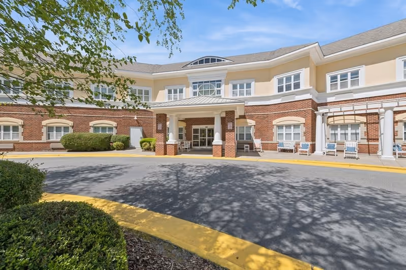 Front exterior view of Fox Hollow Senior Living Community building with a covered entrance, brick and beige facade, multiple windows, bushes, and a circular driveway with yellow curbs under a clear blue sky.