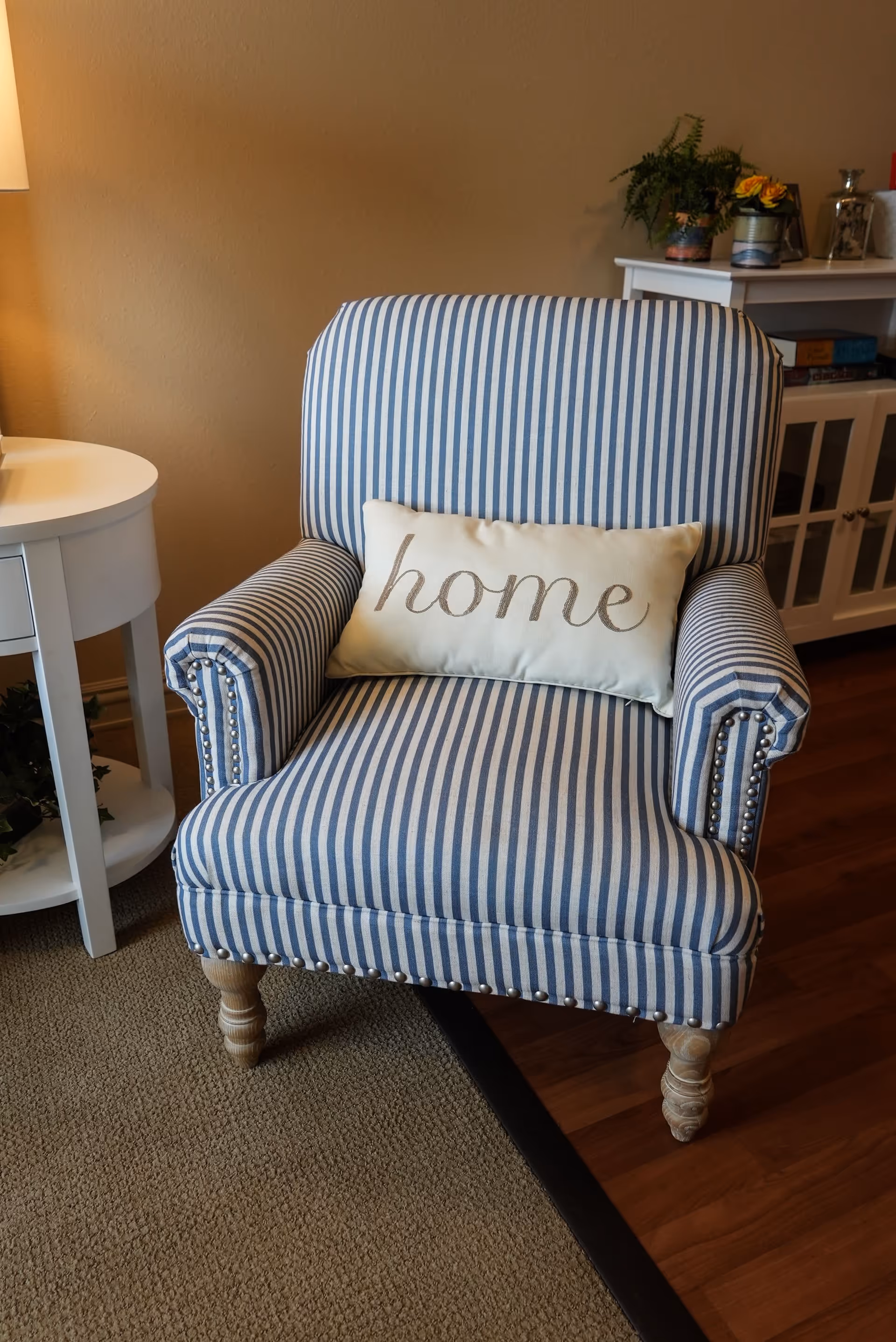 A blue and white striped upholstered armchair with wooden legs and nailhead trim, featuring a rectangular pillow with the word 'home' embroidered on it. The chair is placed on a carpeted floor next to a white round side table with a lamp and a white cabinet with plants and decorative items on top.