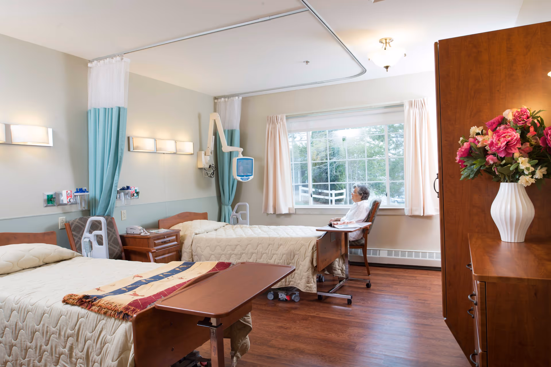 A bright shared care room with two hospital-style beds, a seated resident by the window, medical equipment, and a vase of flowers on a dresser.