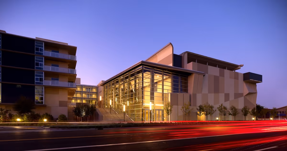 Modern building exterior at dusk with large glass windows and illuminated interior. The building has a multi-story section with balconies on the left and a large angular structure on the right. Trees and shrubs line the front, and red light trails from passing vehicles are visible on the road in front.