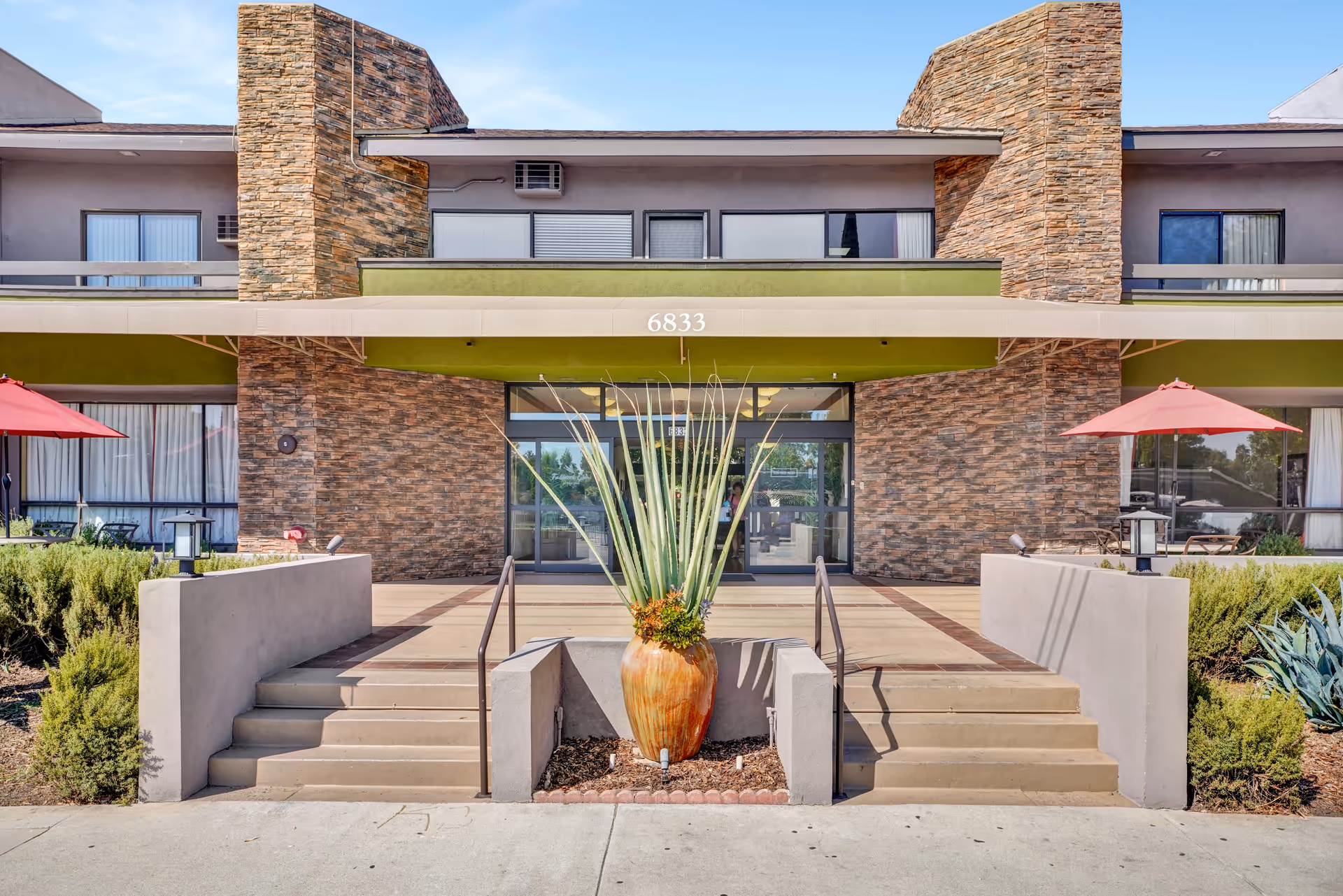 Front entrance of a senior living building with steps, a large potted plant, and the address '6833' above the doorway.