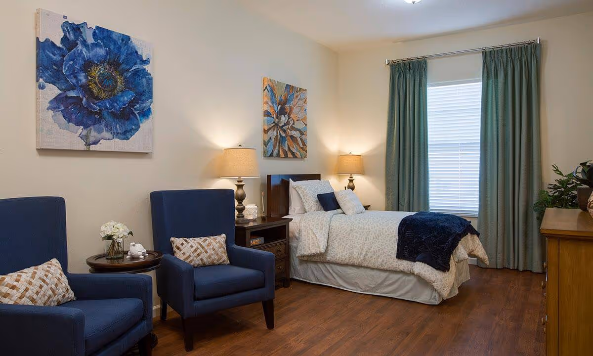 A cozy bedroom with a single bed made up with white and blue bedding, two blue armchairs with patterned pillows, a small round table with a flower vase, two bedside tables with lamps, a wooden dresser, and a window with teal curtains. The walls are decorated with two floral paintings.