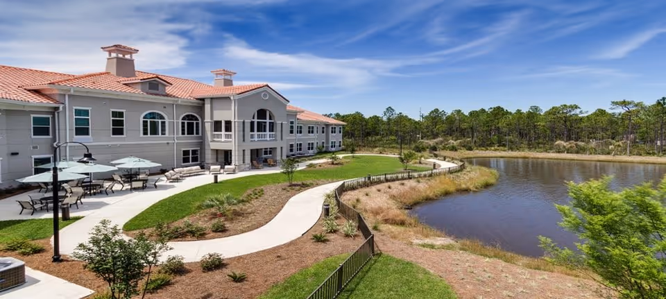 Outdoor view of a senior living facility with a large building featuring a red-tiled roof and multiple windows. In front of the building, there is a paved walkway winding through landscaped gardens with small plants and trees. Several tables with umbrellas and chairs are set up on a patio area. To the right, there is a pond surrounded by grass and trees under a blue sky with some clouds.