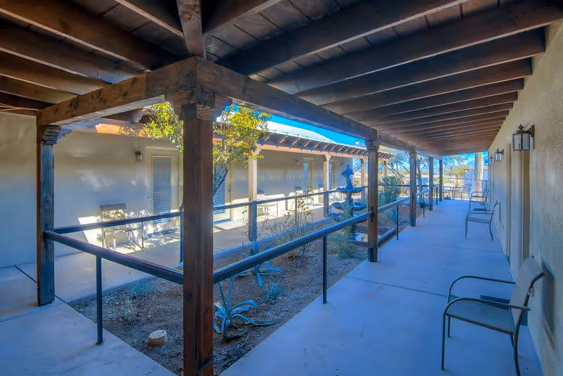 Covered outdoor walkway with wooden beams and railings surrounding a small garden area with plants. Several chairs are placed along the walkway next to the building walls under the roof. The sky is clear and blue in the background.