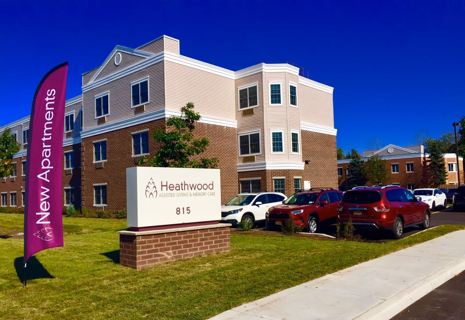Exterior view of the Heathwood Assisted Living & Memory Care building with its front sign, parked cars, and a purple 'New Apartments' flag.