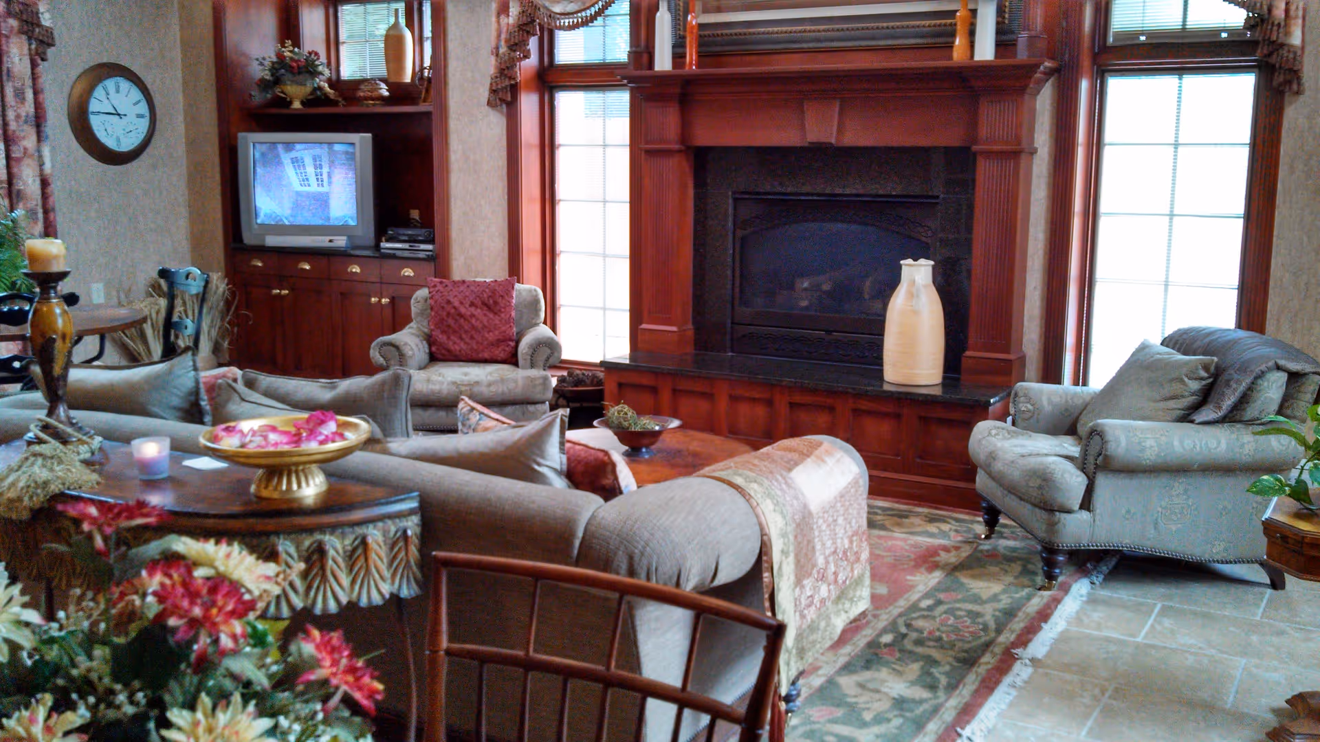 A cozy living room with a large fireplace surrounded by wooden paneling, two armchairs, a sofa with a quilt draped over the back, a wooden coffee table, and a TV set in a built-in wooden cabinet. The room is decorated with floral curtains, a wall clock, and various decorative items including flowers and candles.