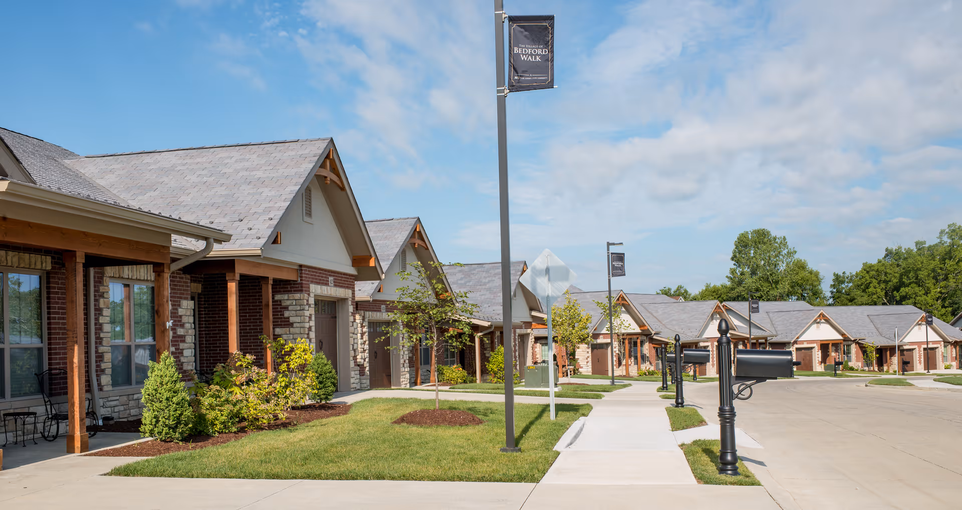 A row of single-story residential buildings with brick and stone facades, each with a small porch and a garage. There are well-maintained lawns, young trees, and black mailboxes along a curved sidewalk under a partly cloudy sky. A street lamp displays a banner that reads 'The Village of Bedford Walk'.
