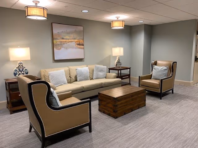 A cozy living room area with a beige sofa adorned with three white pillows, two tan armchairs with light blue pillows, a wooden coffee table, two side tables each with a decorative lamp, and a landscape painting hanging on the wall. The room has soft lighting from ceiling fixtures and table lamps, with neutral-colored walls and carpeted flooring.