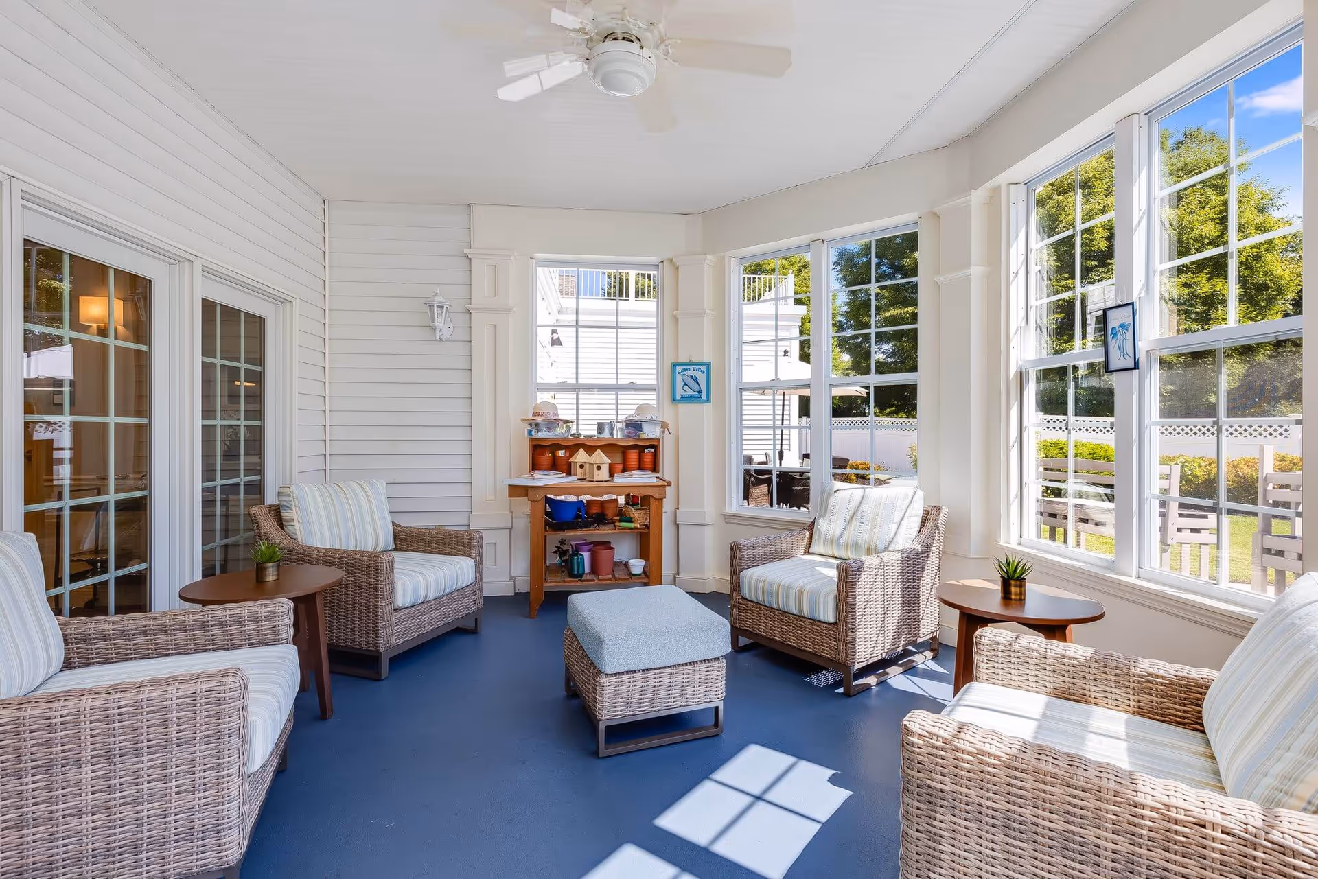 A bright sunroom with large windows showing an outdoor garden area. The room features four wicker armchairs with striped cushions, two small round wooden tables with small potted plants, a wicker ottoman with a blue cushion, and a wooden shelf with various small pots and decorative items. The floor is blue, and the walls are white with paneling. A ceiling fan is mounted on the white ceiling.