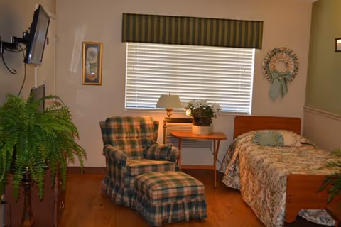 A cozy room with a single bed covered in a floral bedspread, a matching plaid armchair with an ottoman, a wooden side table with a lamp and a potted plant, a large window with blinds and a green valance, a wall decoration with a wreath, and a flat-screen TV mounted on the wall. There is also a large green fern plant on a stand.