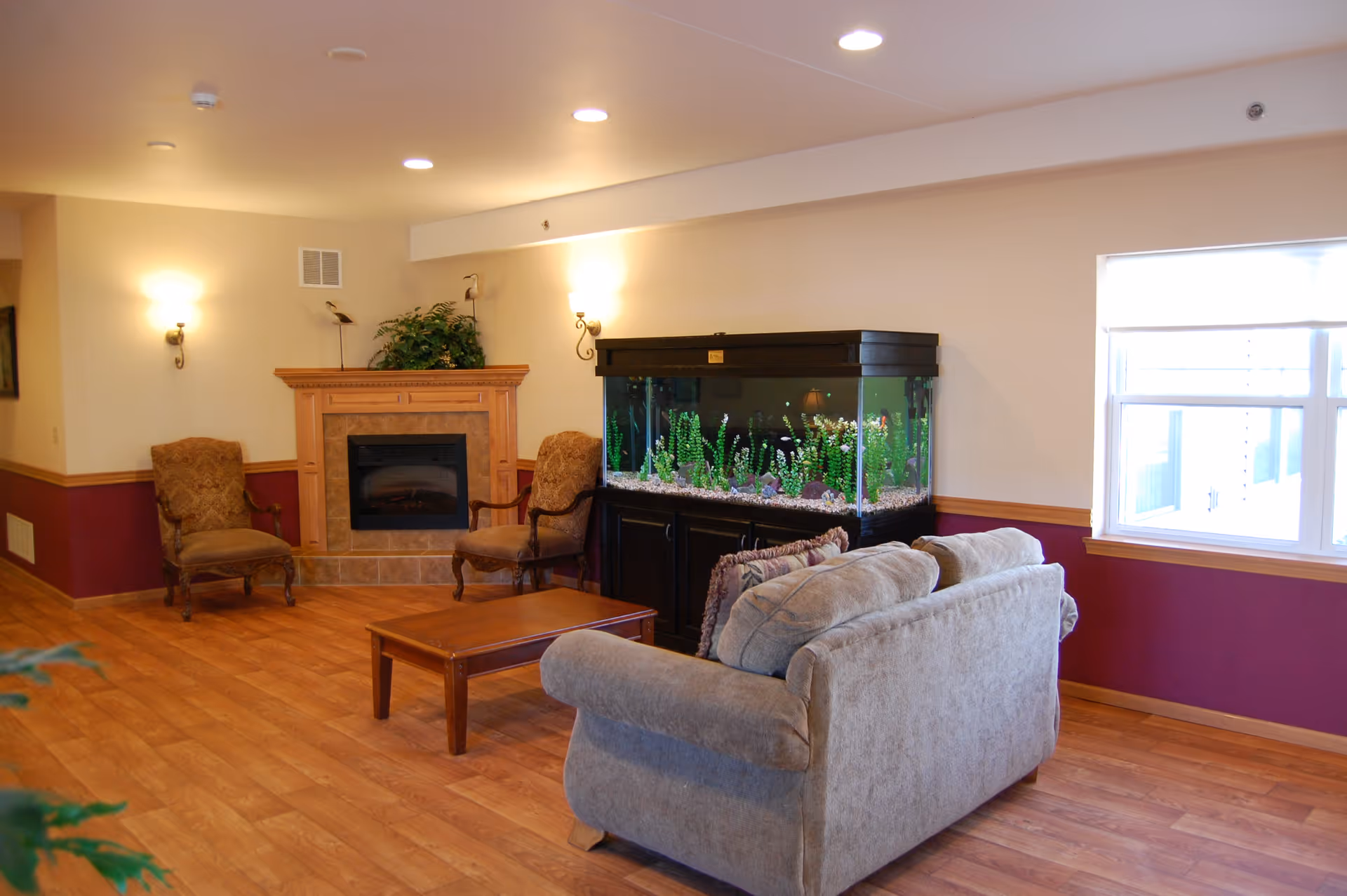 A cozy living room area with a beige couch, two upholstered armchairs, a wooden coffee table, a large fish tank on a black stand, and a fireplace with a wooden mantle. The walls are painted beige with a maroon lower half, and there is a window letting in natural light.