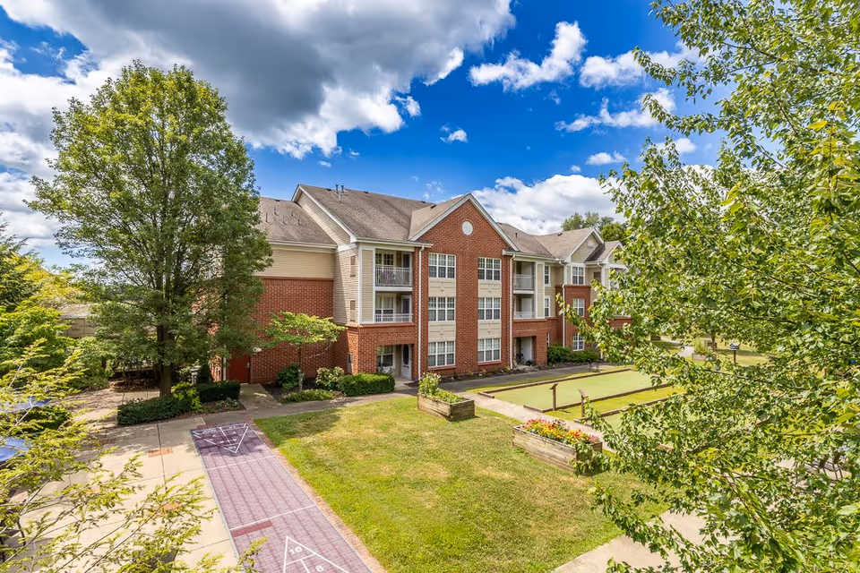 Exterior view of a three-story senior living facility building with red brick and beige siding. The building is surrounded by green trees and a well-maintained lawn with a shuffleboard court and raised flower beds. The sky is partly cloudy with blue patches visible.
