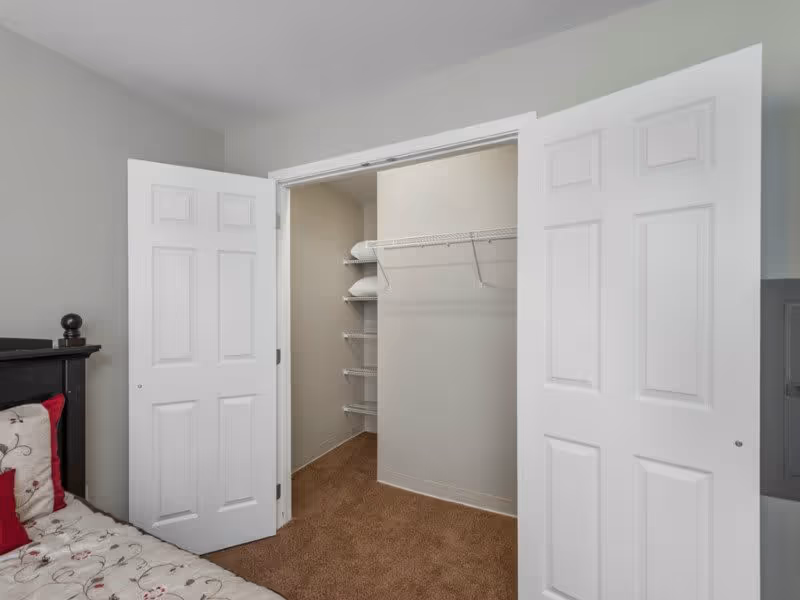 Interior view of a bedroom closet with white double doors open, showing empty wire shelving and a hanging rod. Part of a bed with a floral bedspread and red pillows is visible on the left side.