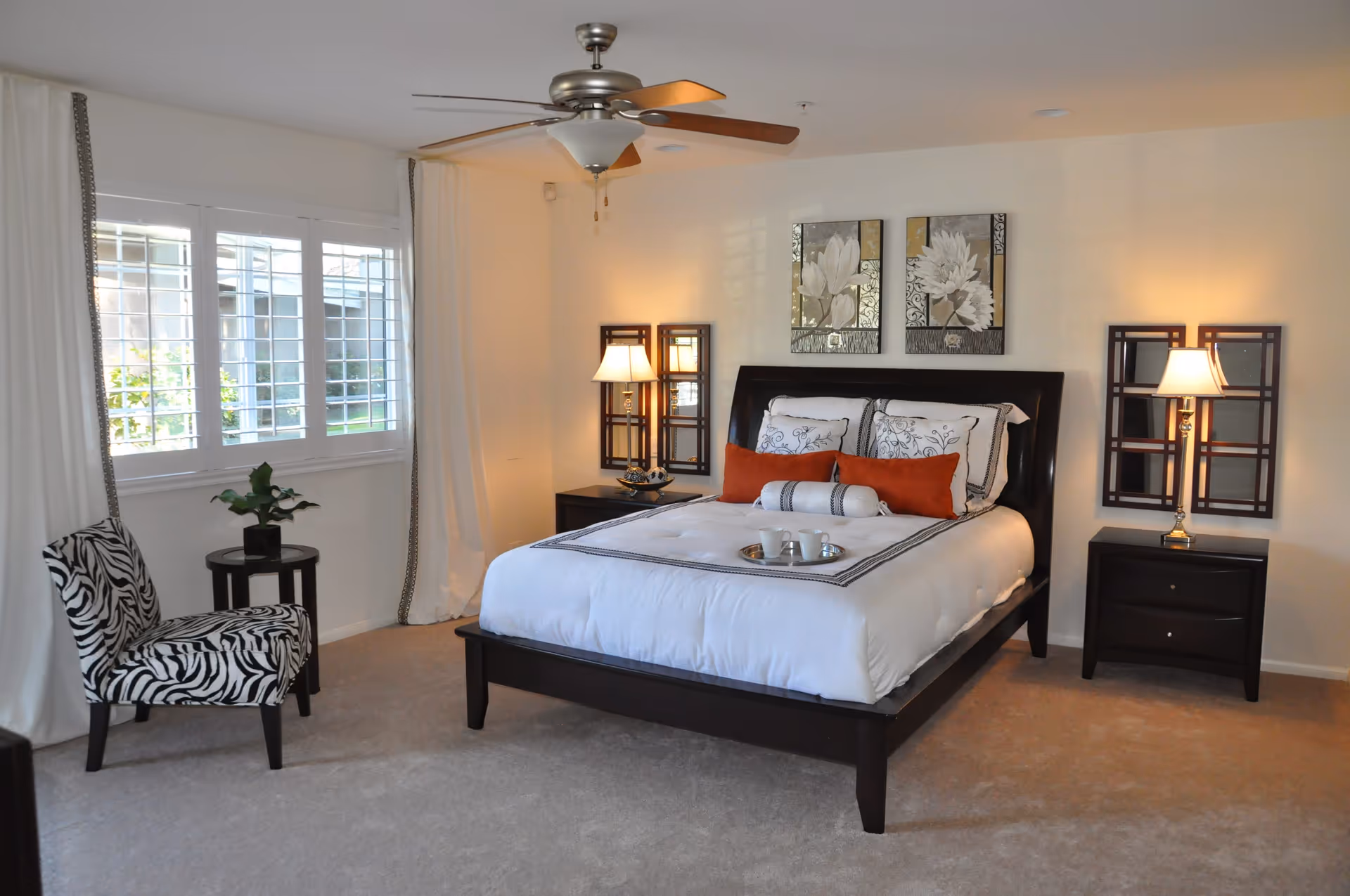 A neatly arranged bedroom with a dark wooden bed frame, white bedding accented with orange and patterned pillows, two matching nightstands with lamps, a ceiling fan, a zebra-patterned chair, a small round table with a plant, and two floral paintings on the wall.