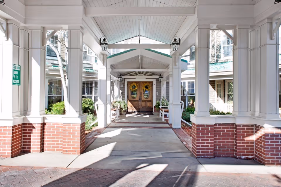 Covered entrance walkway leading to double wooden doors decorated with sunflower wreaths at Sunrise at Orchard facility. The walkway is supported by white columns with brick bases, and there are benches and greenery on either side.