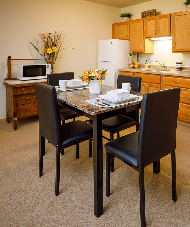 A small dining area in a senior living facility kitchen with a square marble-top table set for four. The table has black chairs around it and a floral centerpiece. In the background, there is a white refrigerator, wooden cabinets, a sink, and a microwave on a wooden side table with decorative plants and flowers.