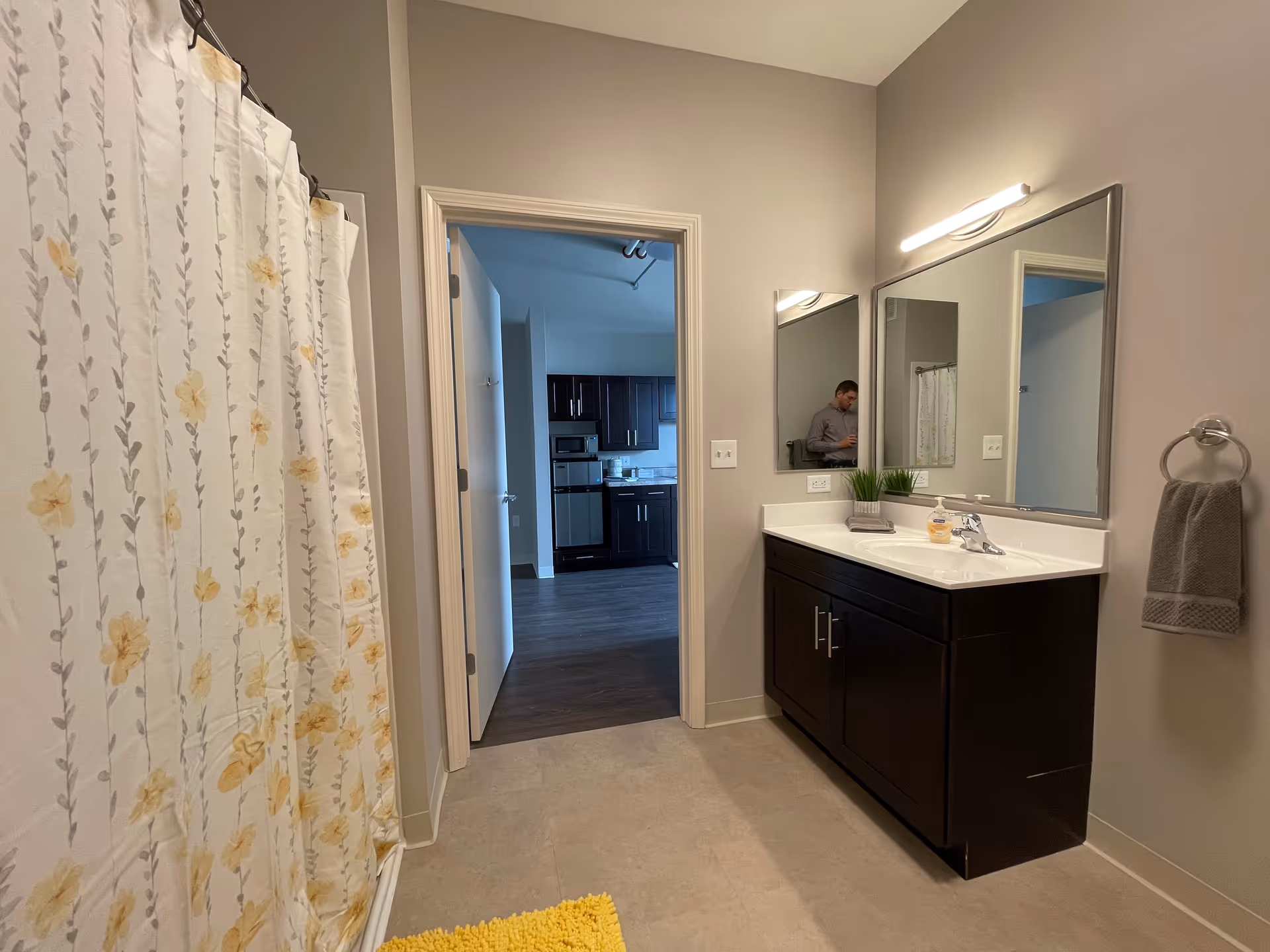 A bathroom with a shower curtain featuring a floral pattern on the left, a dark wood vanity with a white countertop and sink on the right, a large mirror above the vanity, and a towel ring with a gray towel. The bathroom opens into a kitchen area with dark cabinets and stainless steel appliances. A person is partially visible in the reflection of the mirror.