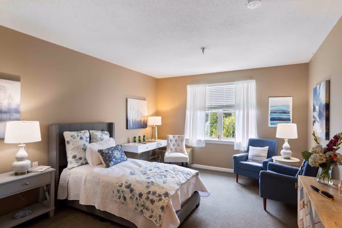 A well-lit bedroom with a single bed featuring floral bedding, two bedside tables with lamps, a white desk with a chair, two blue armchairs with a small round table between them, a window with white curtains, and framed artwork on the walls.