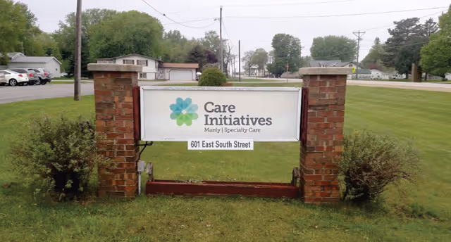 A lawn sign for Care Initiatives Manly Specialty Care reading "601 East South Street" mounted between brick pillars on a grassy lawn.