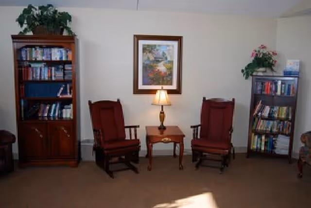 A sitting area with two red upholstered wooden armchairs flanking a small wooden side table with a lamp, bookshelves on either side, and a framed picture on the wall.