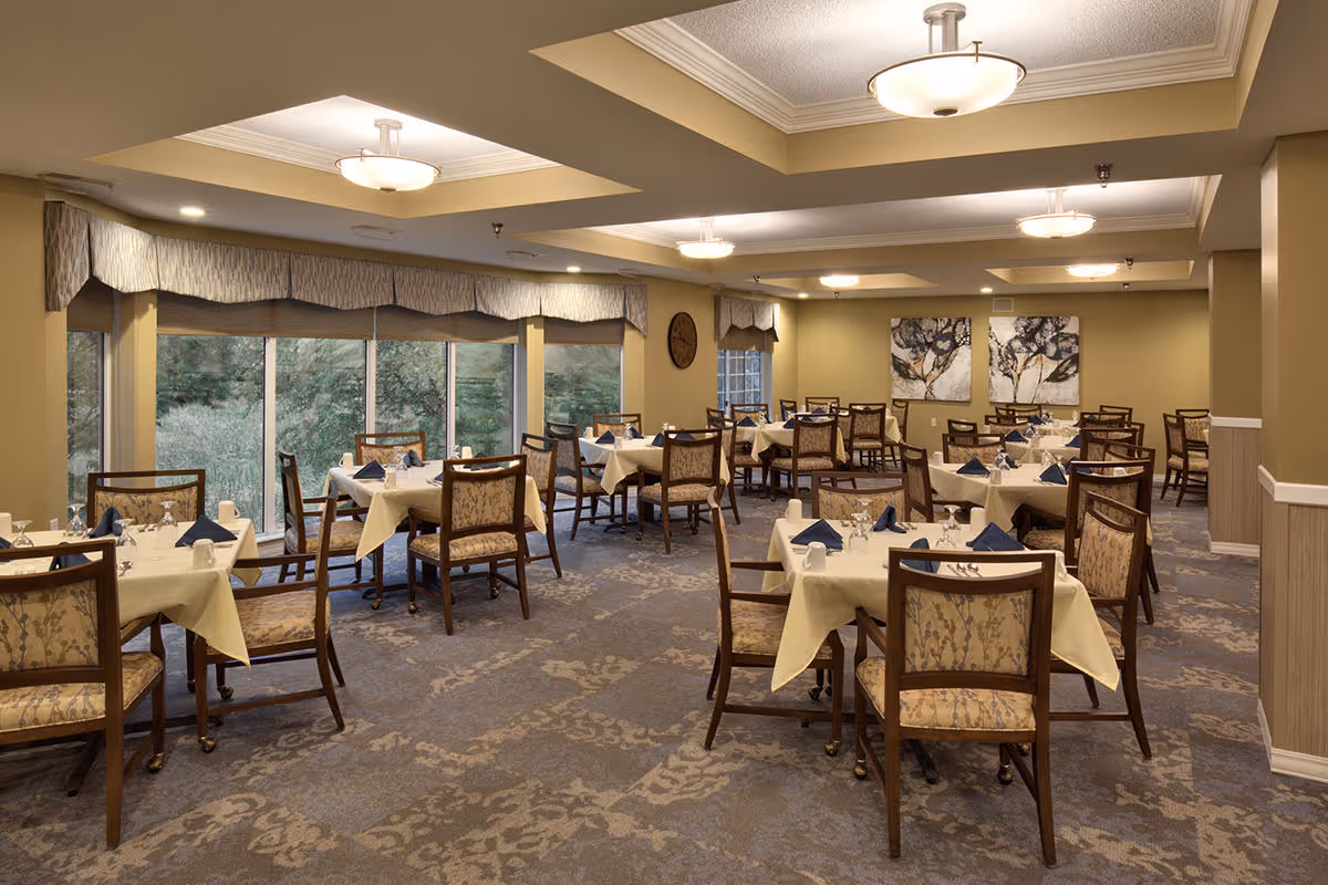 A spacious dining room with multiple tables covered in white tablecloths, each set with blue napkins, glasses, and silverware. The room has large windows with valances, allowing natural light to brighten the space. The walls are painted a warm beige color and decorated with two large floral paintings. The ceiling features recessed lighting and modern light fixtures.