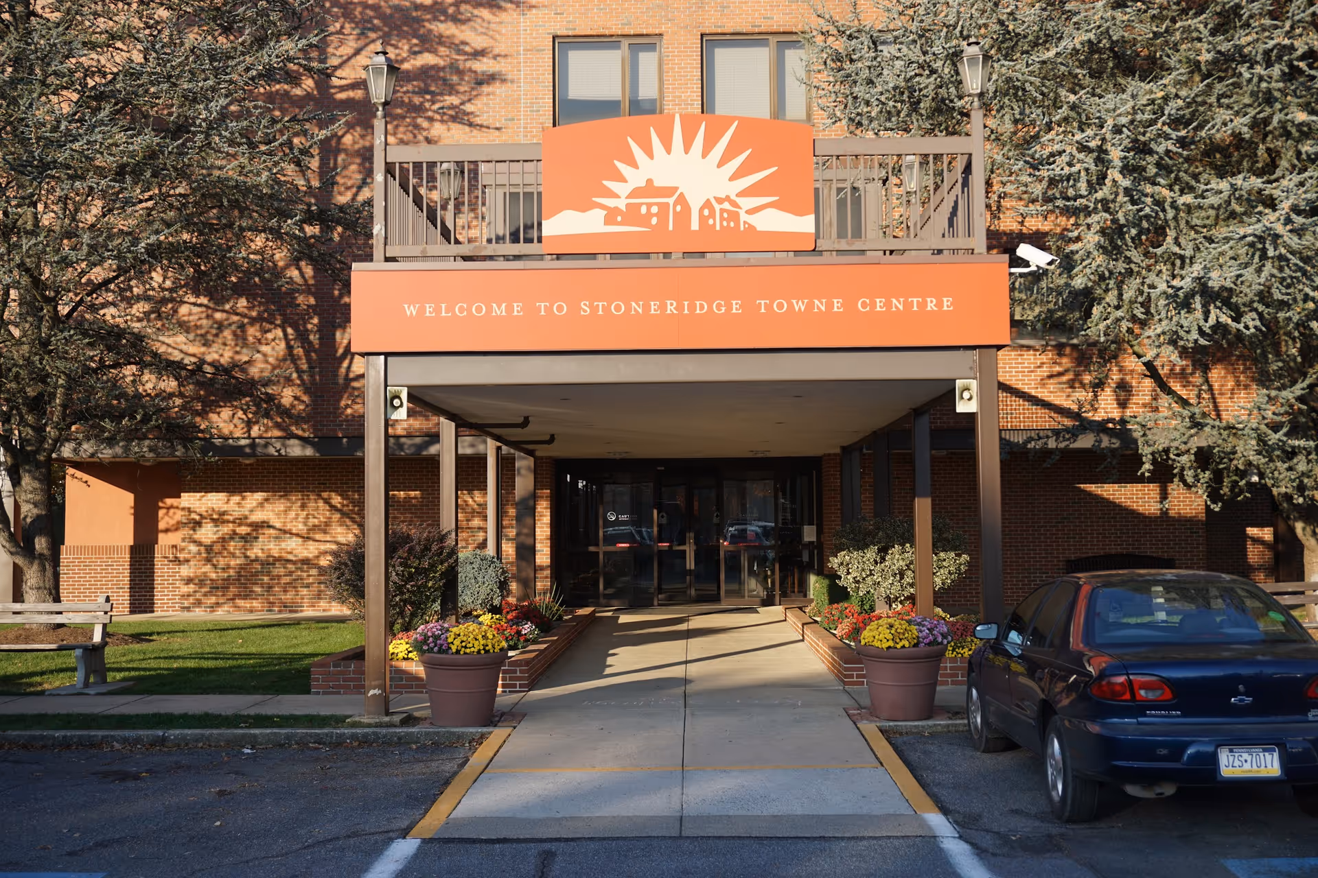 Entrance to a brick building with a covered walkway and a sign above that reads 'Welcome to Stoneridge Towne Centre'. There are flower pots with colorful flowers on either side of the walkway and a parked car to the right. Trees and a bench are visible on the left side.