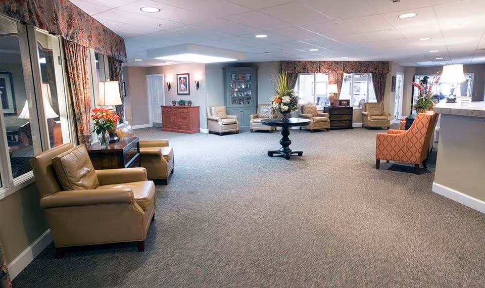 A spacious and well-lit common area in a senior living facility with multiple comfortable armchairs arranged along the walls, a round table with a floral centerpiece in the middle, large windows with floral curtains, and soft carpeting. The room has a warm and inviting atmosphere with lamps and decorative plants.