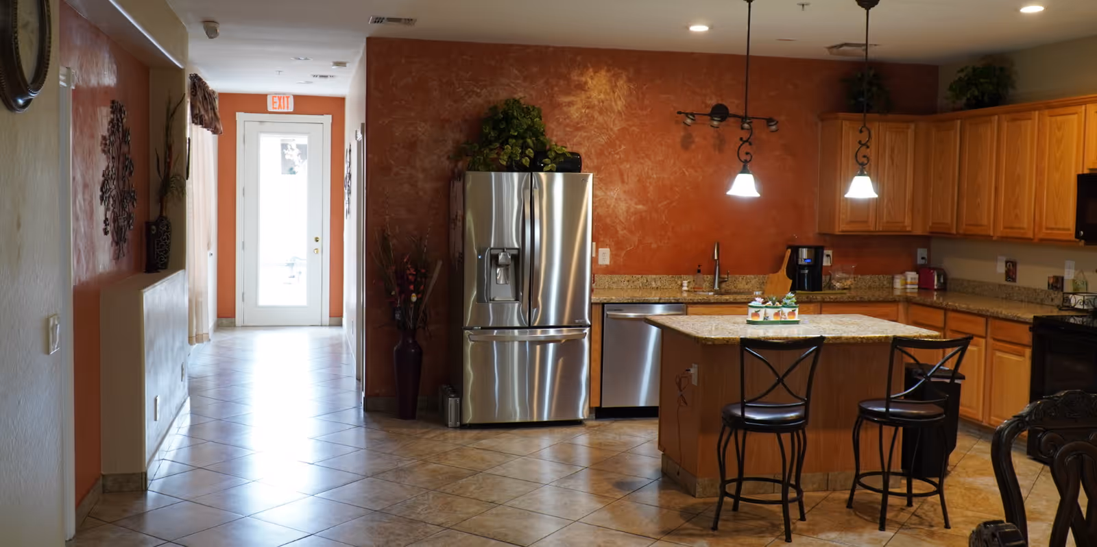 Interior view of a kitchen area with wooden cabinets, granite countertops, a stainless steel refrigerator, dishwasher, and a kitchen island with two black metal bar stools. The walls are painted a warm reddish-brown color, and there are two pendant lights hanging above the island. A hallway with a tiled floor leads to a glass door with an exit sign above it.