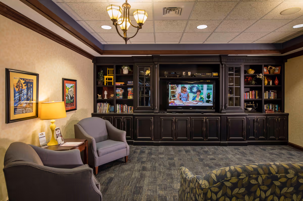 Cozy common living room with upholstered chairs, a patterned sofa, and a flat-screen TV set in dark wood built-in cabinetry with bookshelves and warm lighting.
