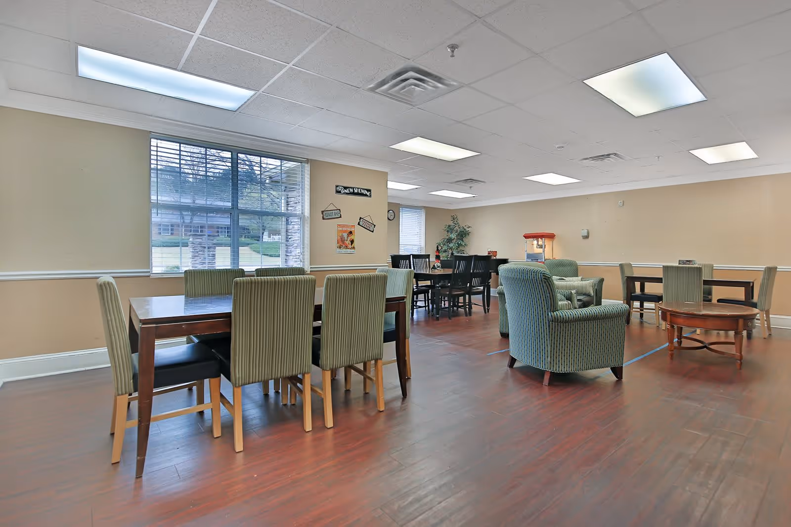 Spacious communal dining and activity room with multiple dining tables, upholstered chairs and armchairs, hardwood floors and large windows.