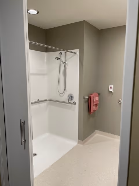 Accessible bathroom with a roll-in shower, grab bars and handheld showerhead, a towel on the wall, and an emergency call button.