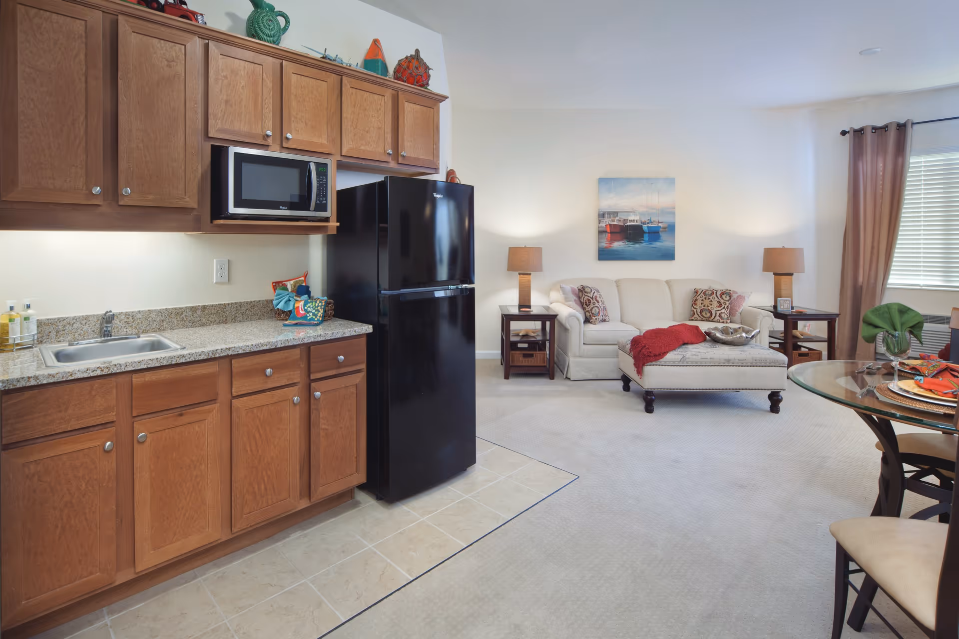 Open-plan kitchen and living area with wooden cabinets and granite countertop, a black refrigerator, beige sofa and ottoman, lamps, and a glass dining table.