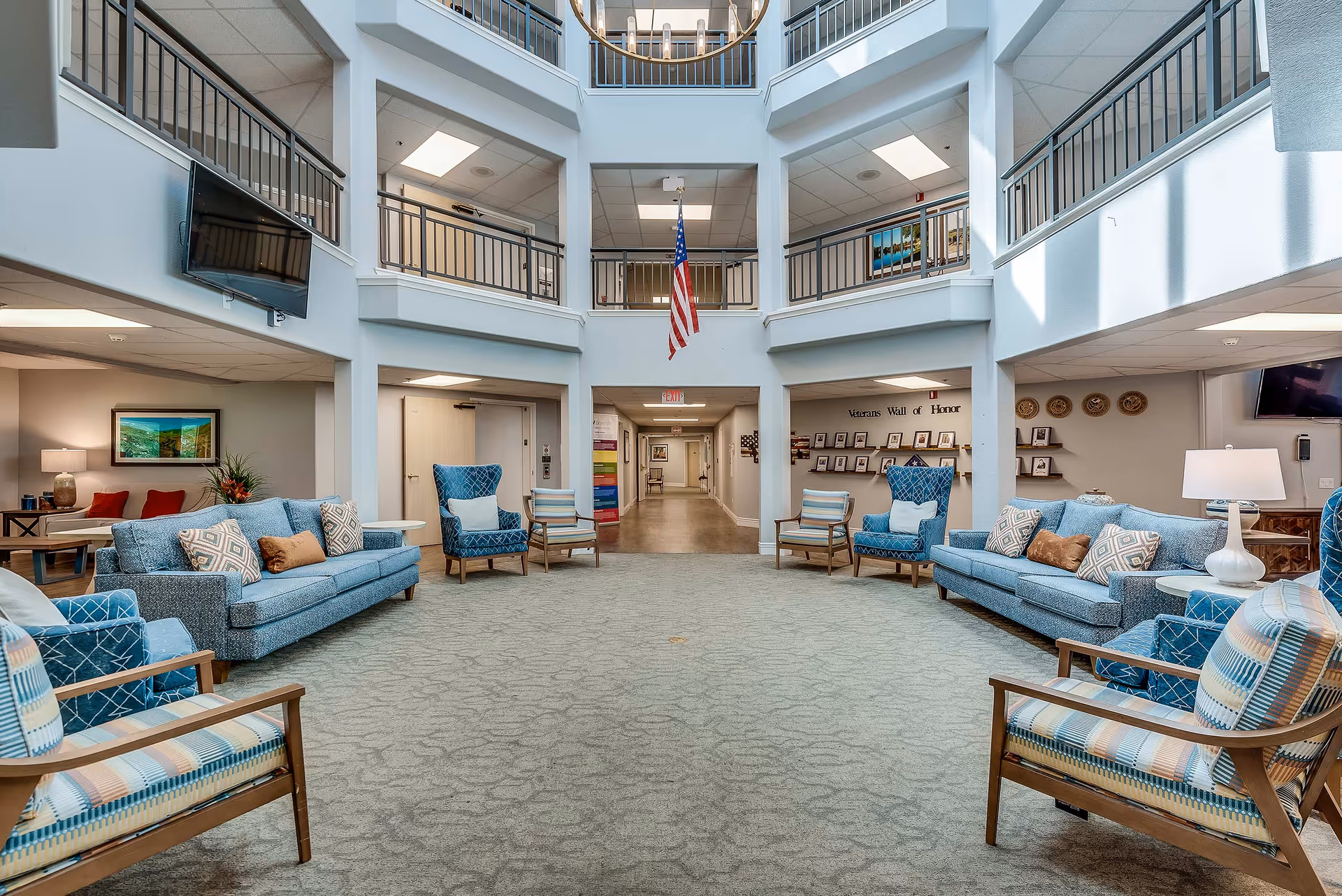 Spacious senior living facility common area with blue upholstered sofas and armchairs arranged around the room. The area features a high ceiling with a second-floor balcony overlooking the space. An American flag hangs in the center above a hallway entrance. The walls display framed pictures and a 'Veterans Wall of Honor'. The room is well-lit with natural light and ceiling lights.