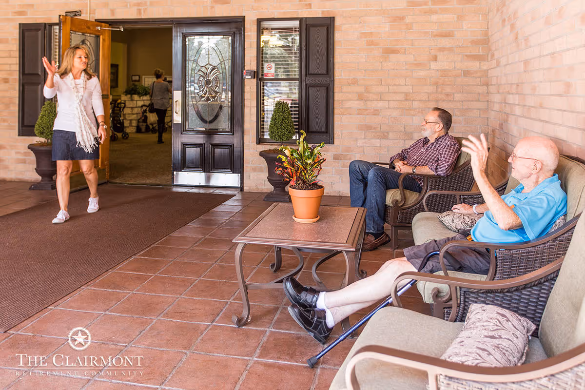Two elderly men sitting on cushioned wicker chairs on a tiled patio, waving and greeting a woman who is walking out of a building entrance with glass and wooden doors. There is a small table with a potted plant between the men and the entrance. The setting appears warm and welcoming with brick walls and decorative plants.