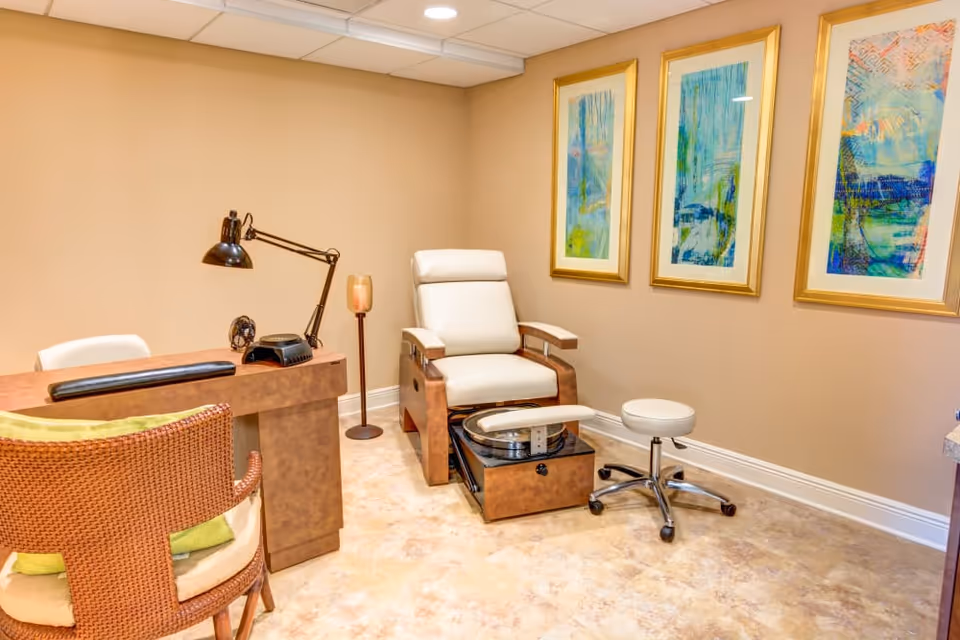 Small treatment room with a desk, reclining spa chair and footbath, rolling stool and framed artwork on beige walls.