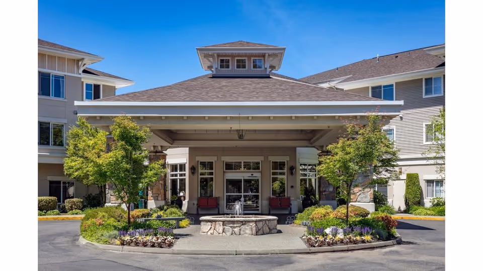 Covered main entrance of a multi-story senior living building with a central stone fountain and landscaped plantings.