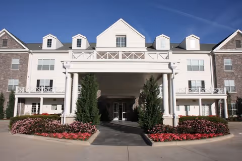 Front exterior view of a multi-story senior living facility named Prestonwood Court with white siding and brick accents, featuring a covered entrance supported by large white columns, landscaped with bushes and flowers.