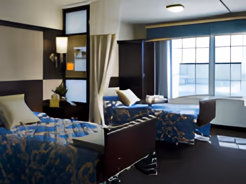 Shared resident bedroom with two hospital-style beds separated by a privacy curtain, bedside tables and a large window.