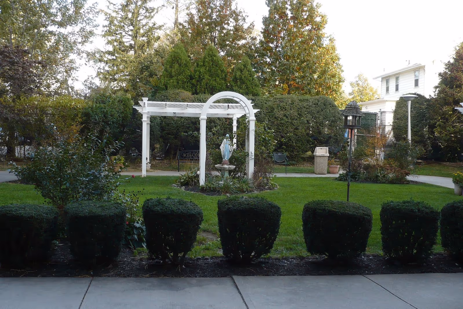 A well-maintained garden area with neatly trimmed bushes in the foreground, a white pergola with an arch, and a statue of a person in the center surrounded by greenery. Trees and shrubs form a natural backdrop, and a white building is visible in the background on the right side.