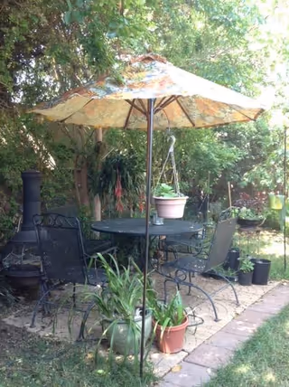 Outdoor patio area with a round metal table and four matching chairs under a large floral-patterned umbrella. Several potted plants are placed around the table, and there is a chiminea fireplace to the left. The area is surrounded by green trees and bushes, creating a shaded garden setting.