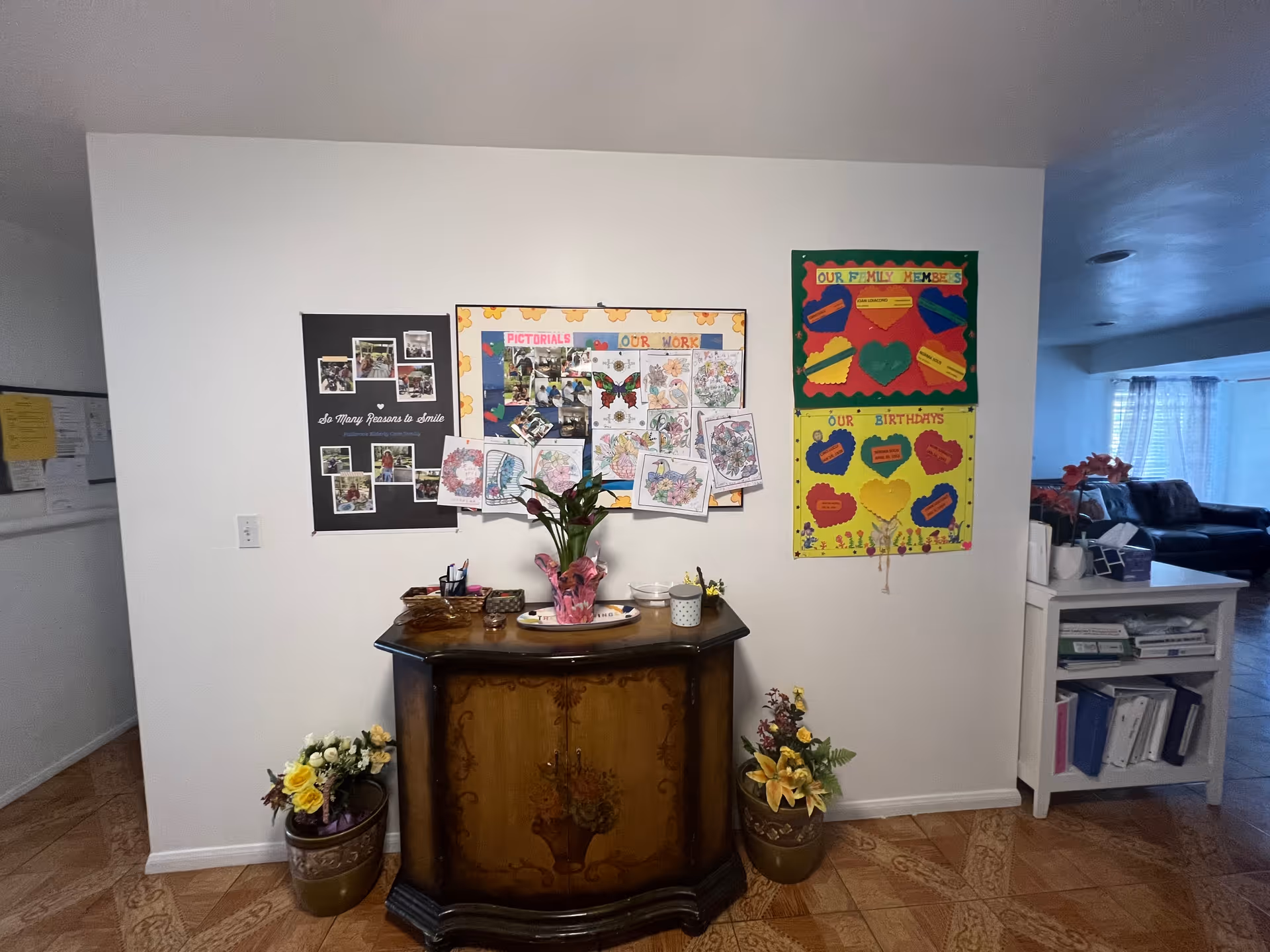 Interior view of a senior living facility showing a decorative wooden cabinet with a flower vase on top, flanked by two flower pots on the floor. Above the cabinet, there are three colorful bulletin boards displaying photos, drawings, and information about family members and birthdays. To the right, part of a living room with a couch and window with curtains is visible.