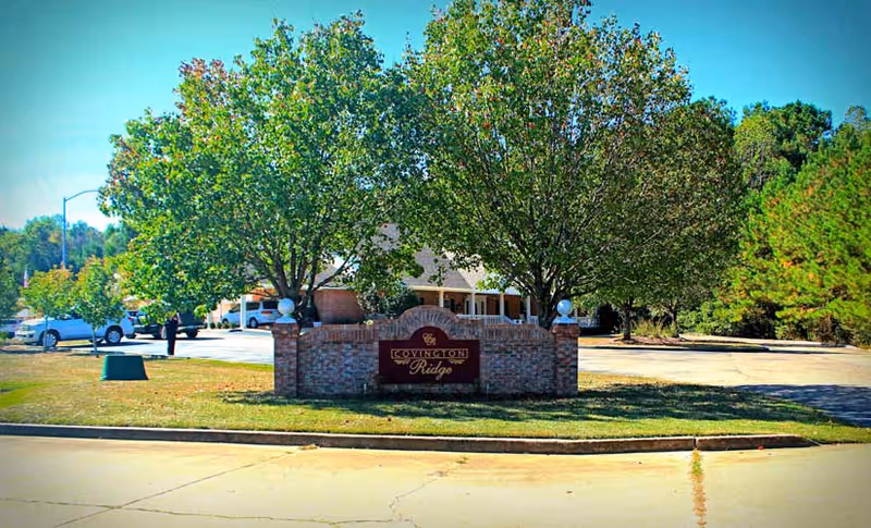 Exterior view of Covington Ridge Retirement Home sign made of brick with two trees behind it, a parking lot with cars, and a building partially visible in the background under a clear blue sky.
